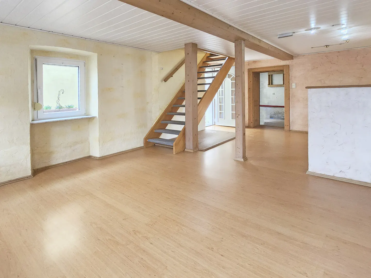 Spacious room with light wood floors, textured yellow walls, and a white paneled ceiling. A wooden staircase leads up, and a window provides natural light.
