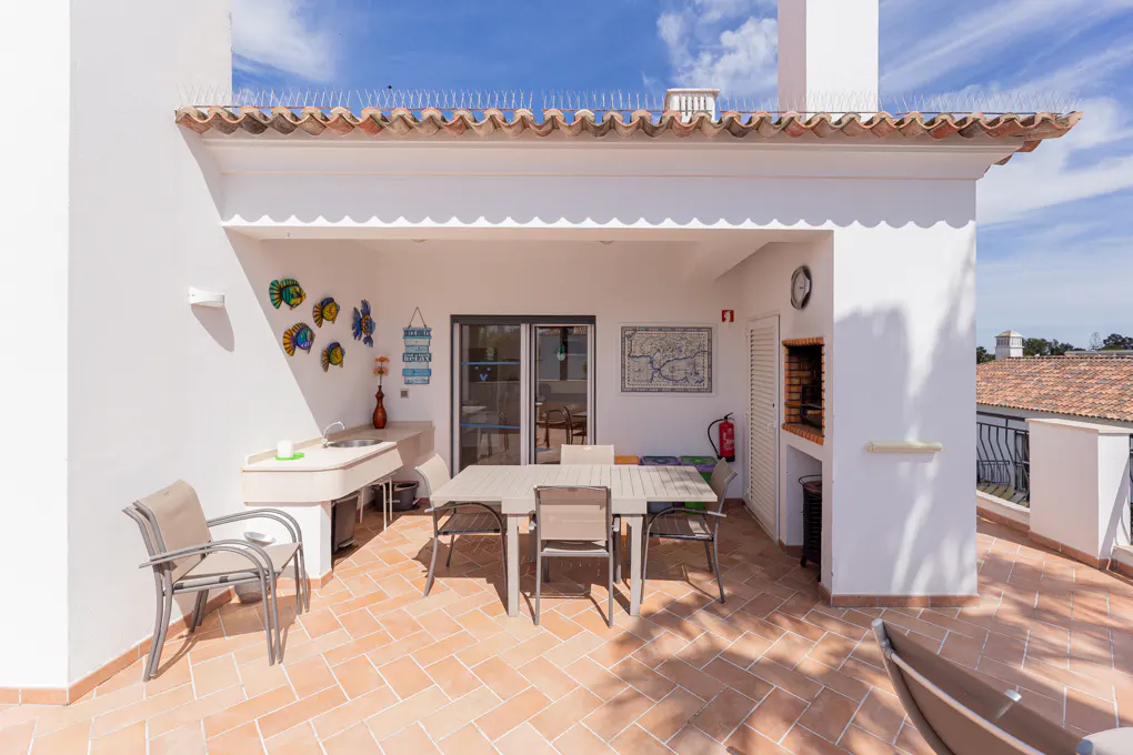 Outdoor patio with white walls, terracotta tile floor, dining table, sink, and built-in grill. Blue sky visible above.