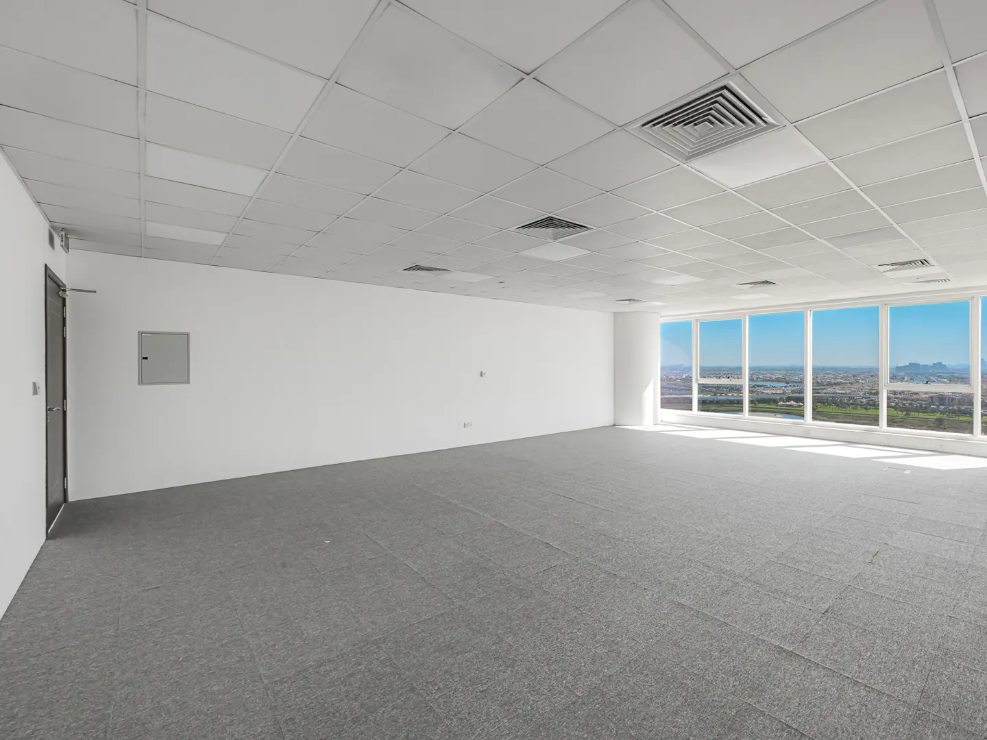 Empty office space with gray carpet, white walls, and a tiled ceiling. Large windows offer a city view.