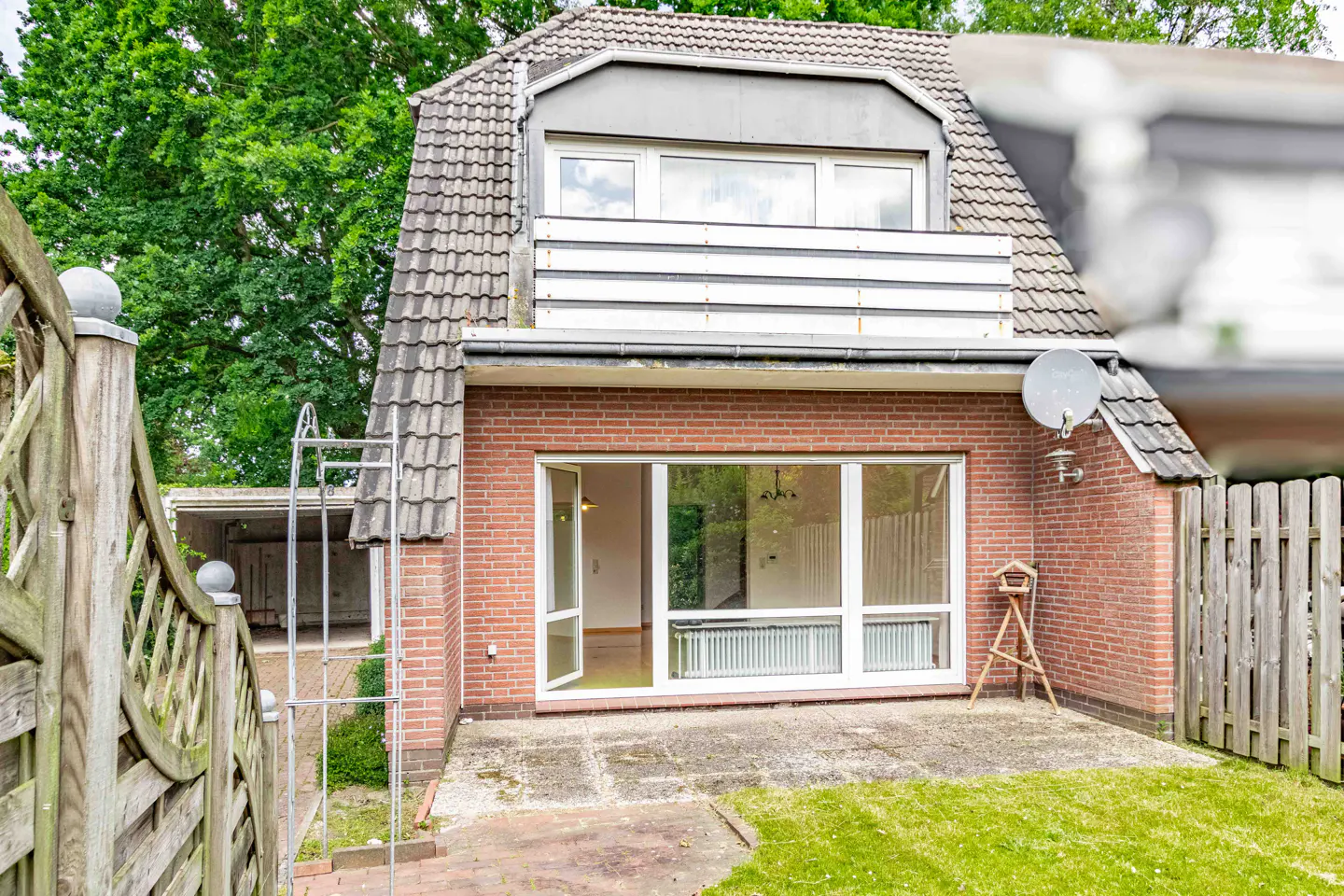 Red brick house exterior with a patio, large glass sliding doors, a balcony, and a wooden fence in a green garden.