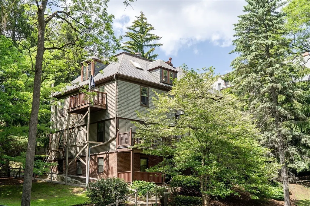 Three-story house with shingle siding, brick base, and wooden balconies, surrounded by lush green trees and landscaping.
