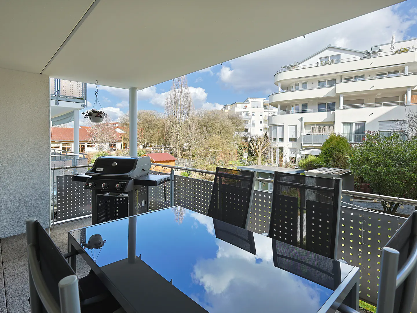 Balcony with a black grill, table, and chairs. The table reflects the sky. White buildings and trees are in the background.