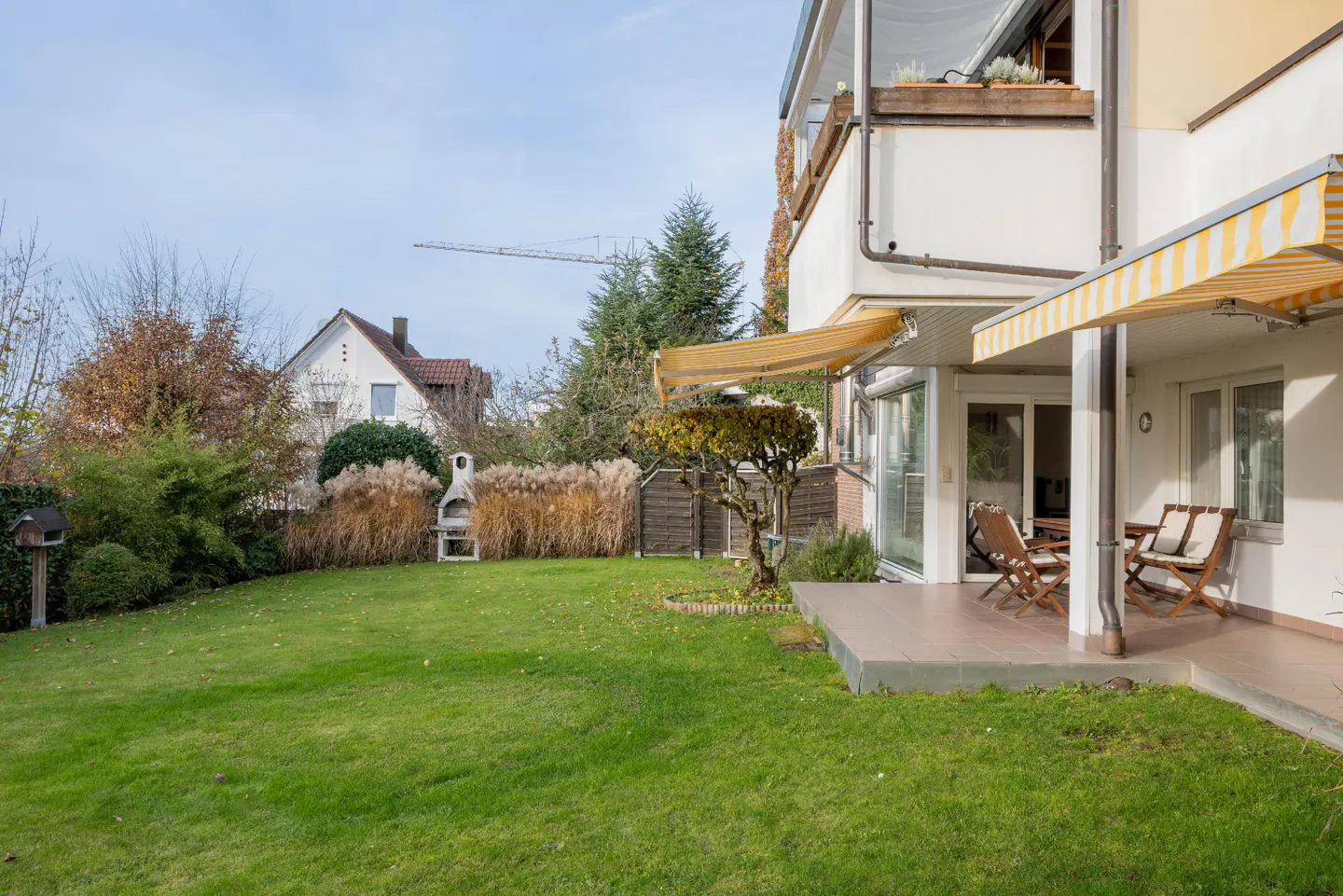 A house with a green lawn, patio with chairs, and a grill in the backyard.