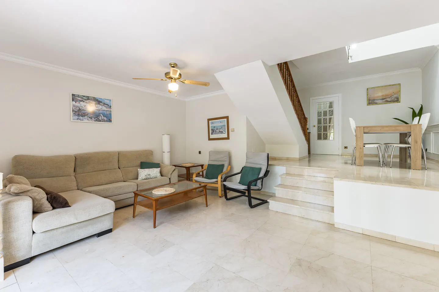 Living room with beige sectional sofa, two chairs, and a wooden table. A staircase leads to a dining area with a table and chairs.