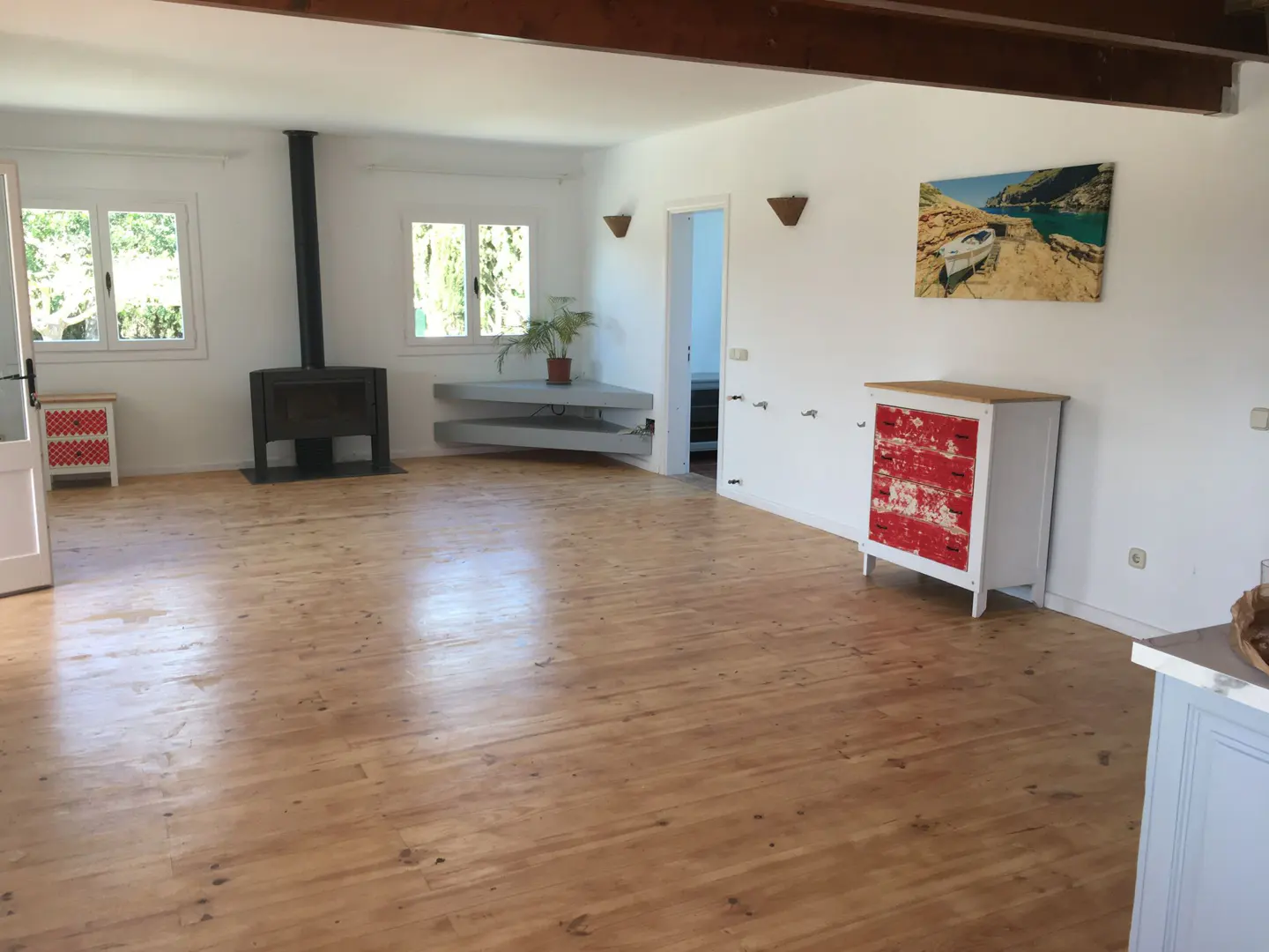 Bright living room with wood floors, white walls, and exposed beams. A black fireplace, red dresser, and boat painting add character.