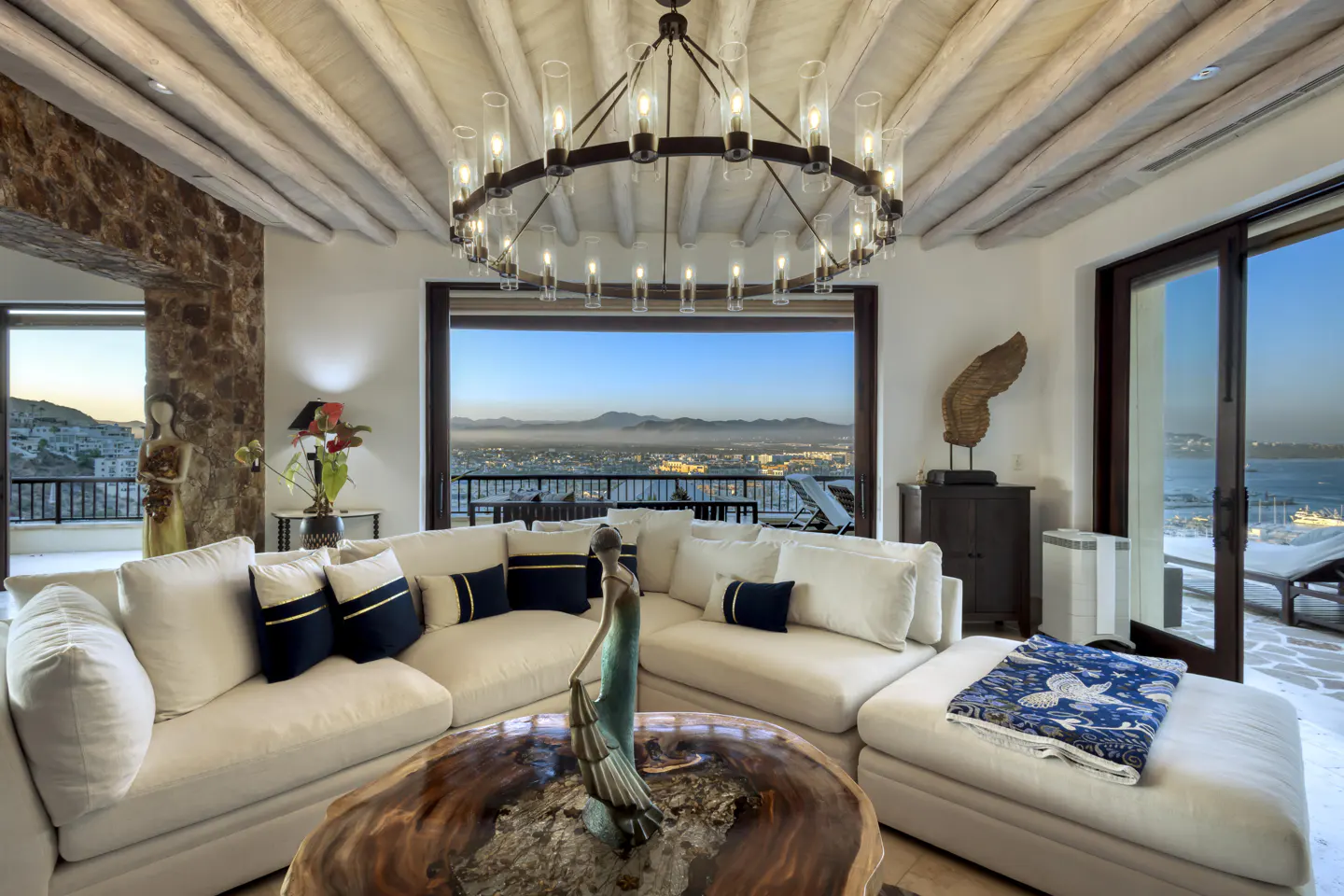 Living room with white sectional sofa, wood coffee table with statue, and city view through large windows. Round chandelier hangs above.