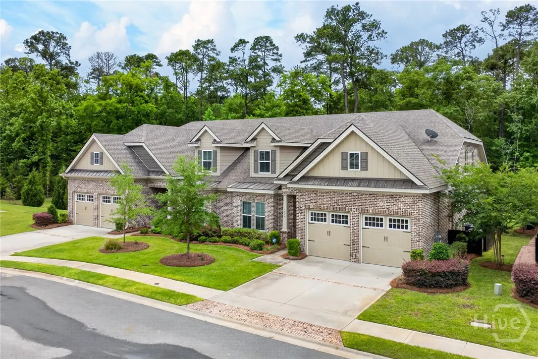 Two-story brick house with a gray roof, tan garage doors, and green lawn. Trees in the background.