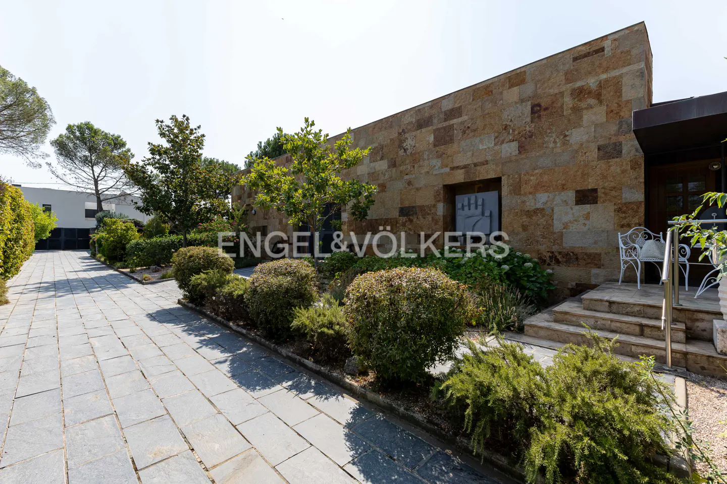 Modern home exterior with stone facade, stone-paved walkway, manicured shrubs, and a small patio with white metal chairs.