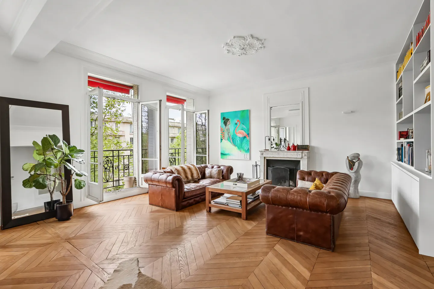 Bright living room with herringbone wood floors, brown leather sofas, and open French doors to a balcony. A fireplace and built-in shelves add charm.