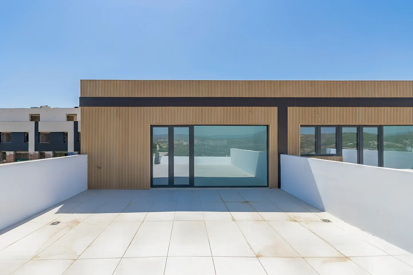 Exterior view of a modern building with a white tiled terrace, white walls, and wood-paneled facade under a clear blue sky.