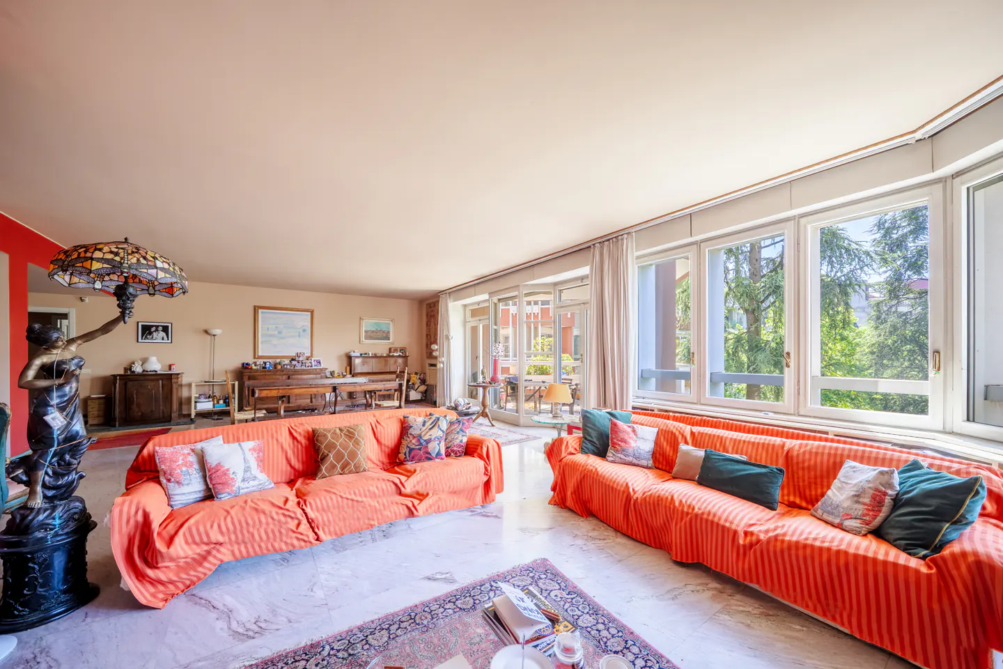 Bright living room with two orange sofas, marble floors, and large windows overlooking greenery. A statue lamp stands near the sofa.