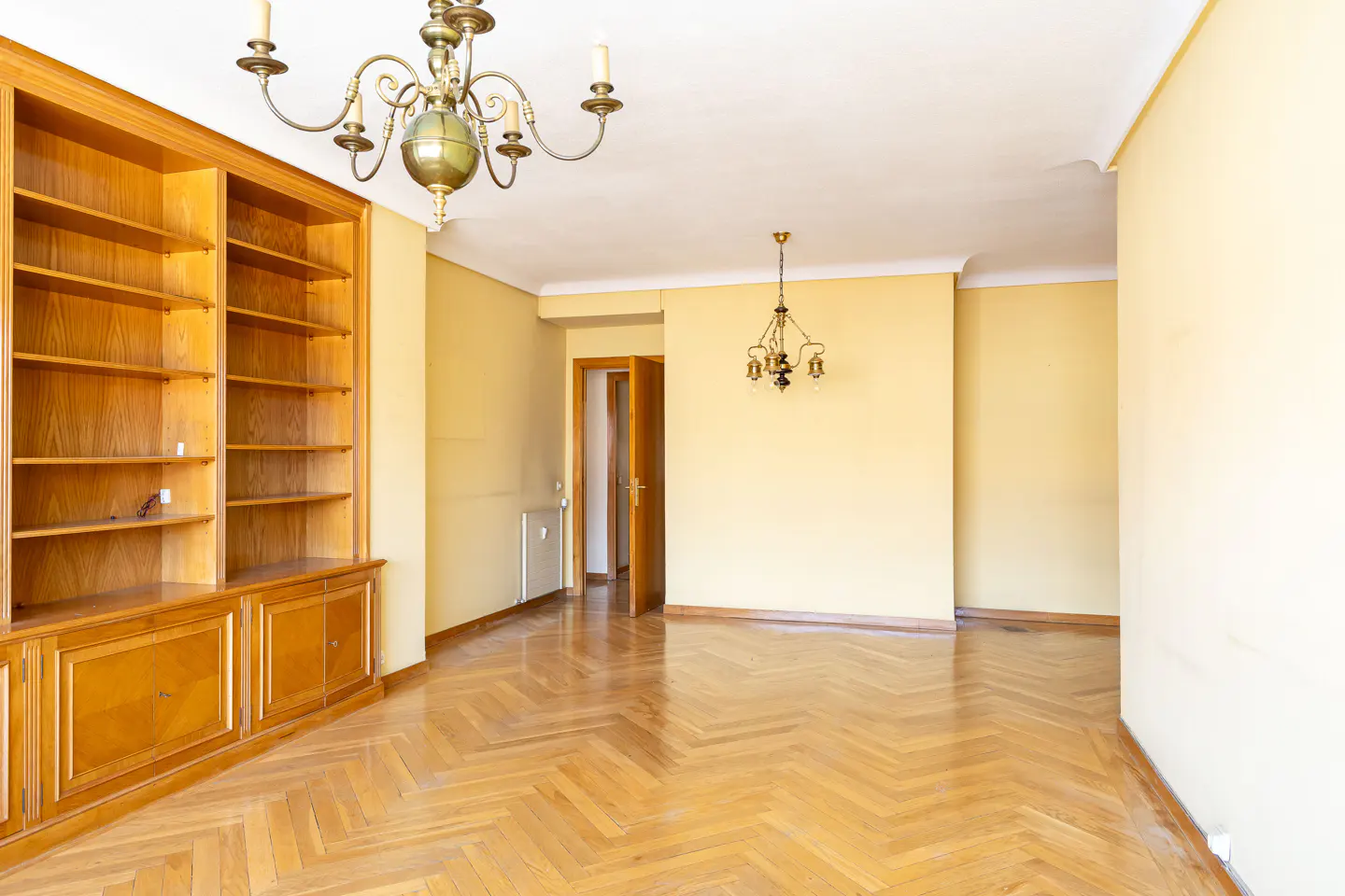 Empty room with herringbone wood floors, yellow walls, and a large wooden bookcase. Two chandeliers hang from the ceiling.