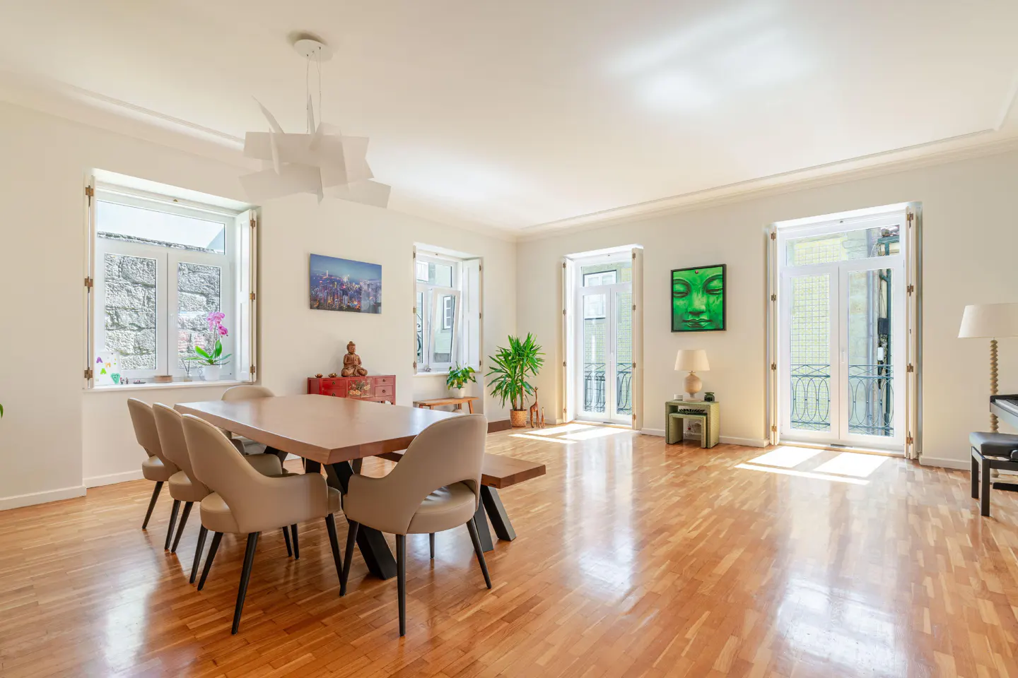 Bright, airy dining room with wood floors, a large table with chairs, and windows. Artwork and plants add color.