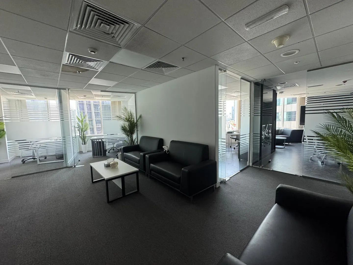 Office space with black leather sofas, a white table, and glass-walled offices with desks and chairs.