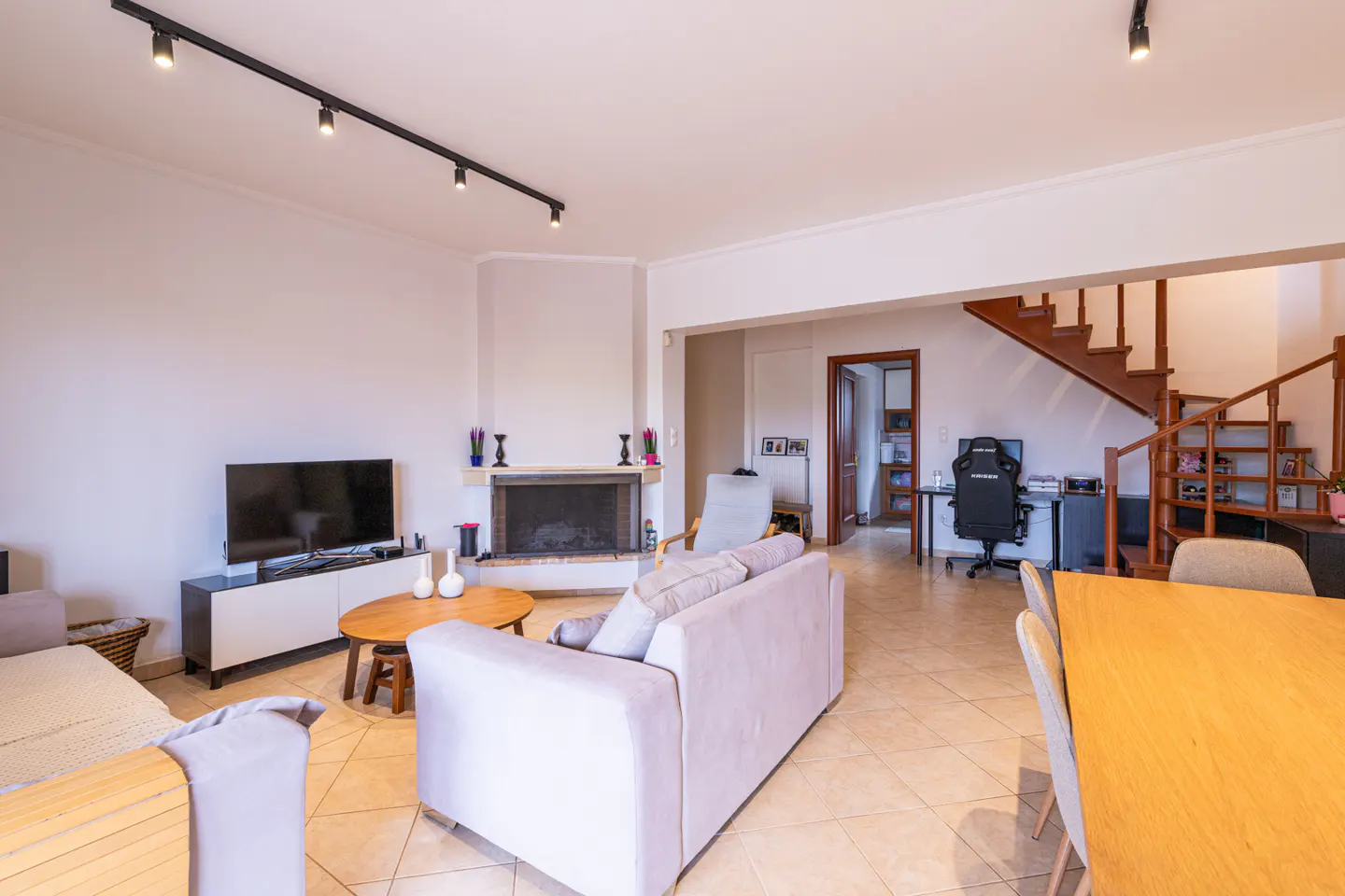 Open-plan living room with light grey sofa, fireplace, TV, dining area, and wooden staircase leading to an upper floor.