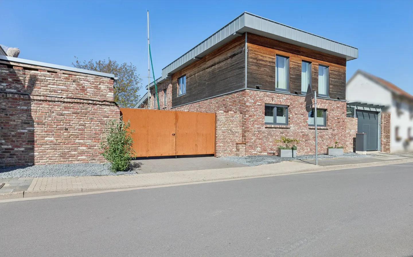 Modern two-story house with brick and wood siding, a metal roof, and a large, rusty metal gate on a sunny day.