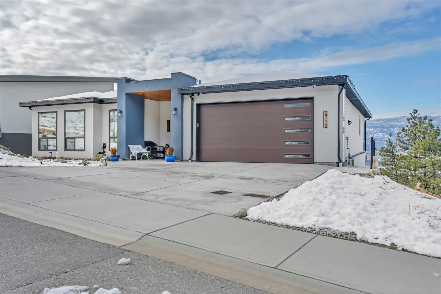 A modern, single-story home with a brown garage door and blue accents on a cloudy day. Snow is visible on the ground.