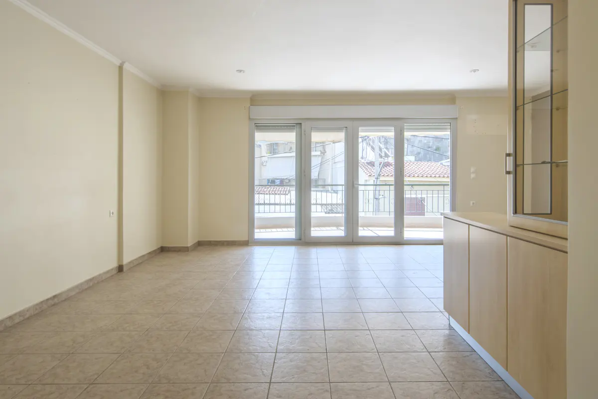 Bright, empty room with beige tile floor, light yellow walls, and glass doors to a balcony. A light wood cabinet stands to the right.