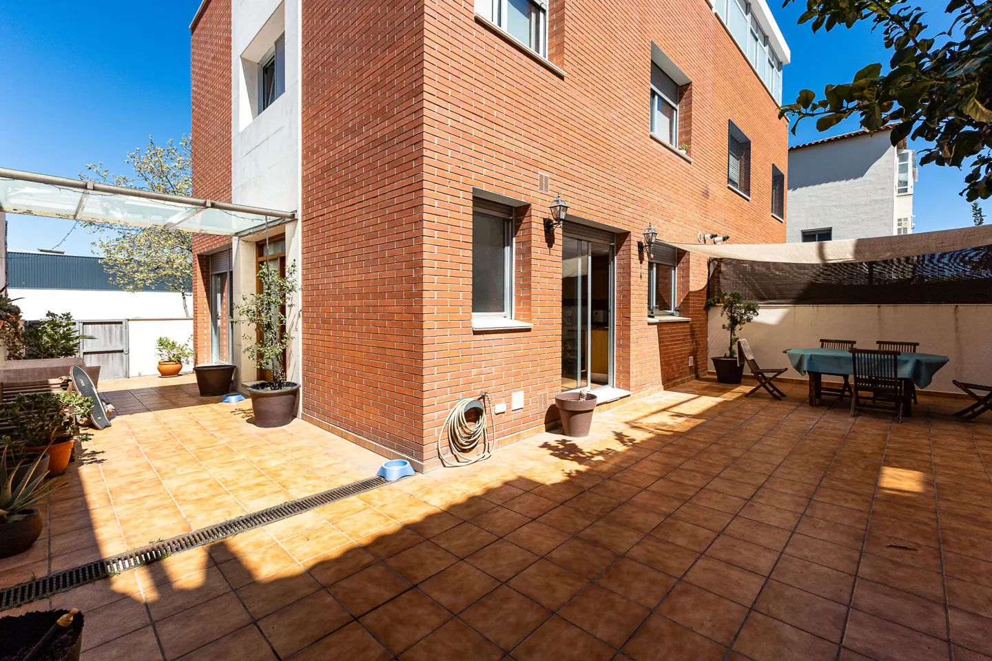 Brick house with a tiled patio featuring a dining table, chairs, and potted plants under a shade sail.