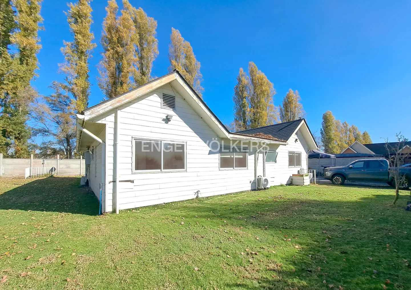 Exterior view of a white, single-story house with a black roof, green lawn, and tall trees under a blue sky.