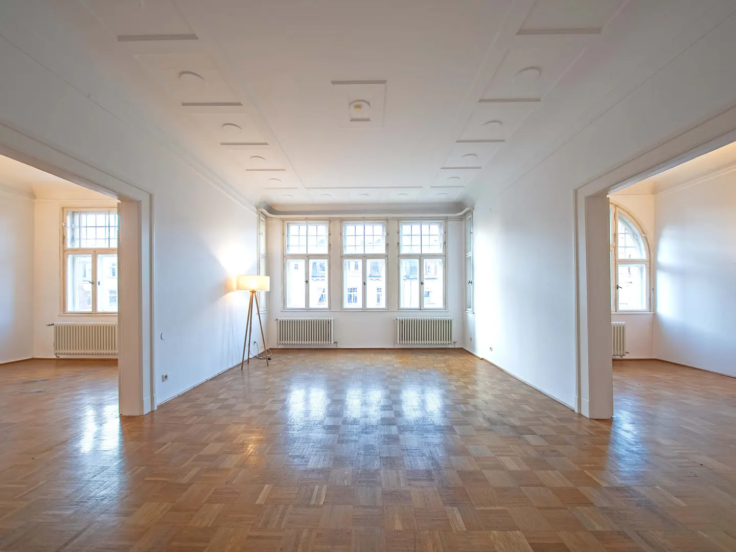 Bright, empty room with parquet floors, white walls, and large windows. A floor lamp stands in the corner. Doorways lead to other rooms.