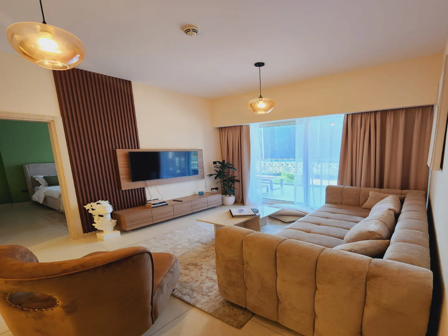 A modern living room with a beige sectional sofa, a flat-screen TV, and a brown accent wall. A bedroom is visible through a doorway.