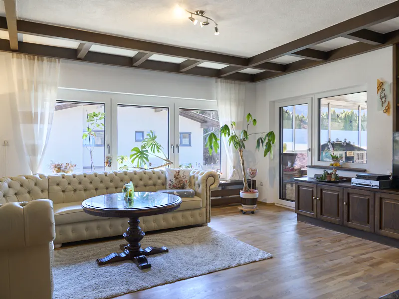 Living room with a beige tufted sofa, round glass table, and wood floors. Large windows offer natural light. A brown wood beam ceiling adds architectural detail.
