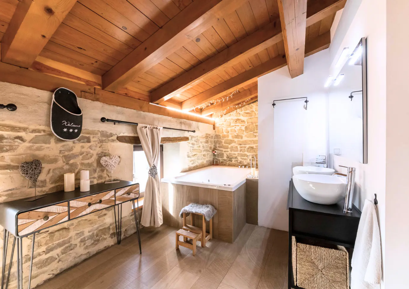 Rustic bathroom with stone walls, wood ceiling, and modern fixtures. A white tub sits near a window with beige curtains. A black console table is on the left.