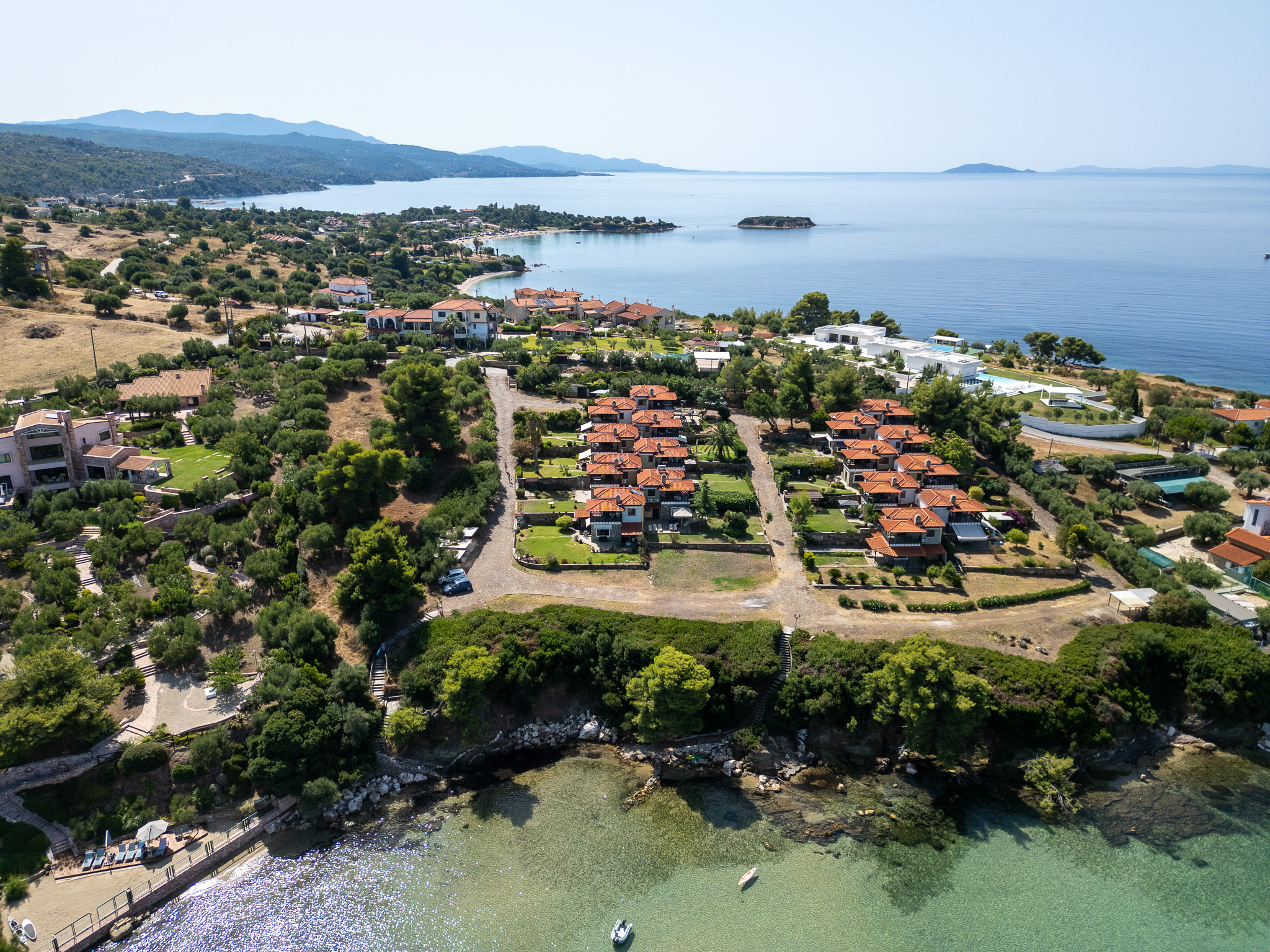 Aerial view of a coastal resort with terracotta-roofed villas, lush green trees, and a clear blue sea in Greece. [AI-generated text]