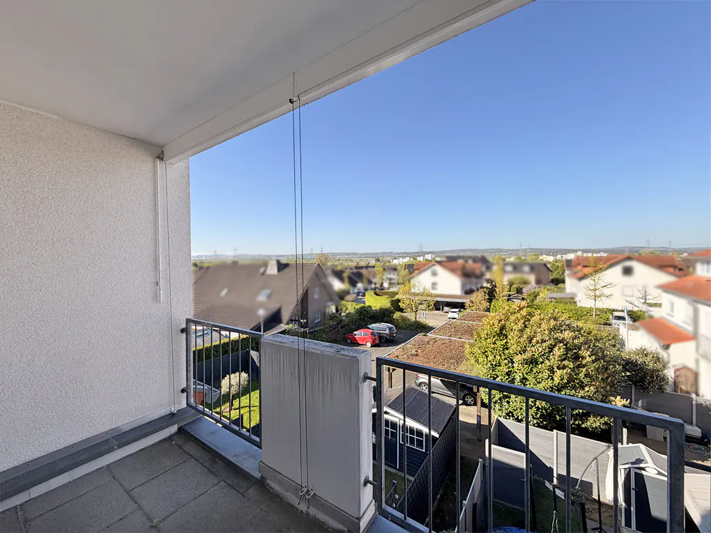 View from a balcony with a gray metal railing overlooking a residential area with houses and trees under a clear blue sky.