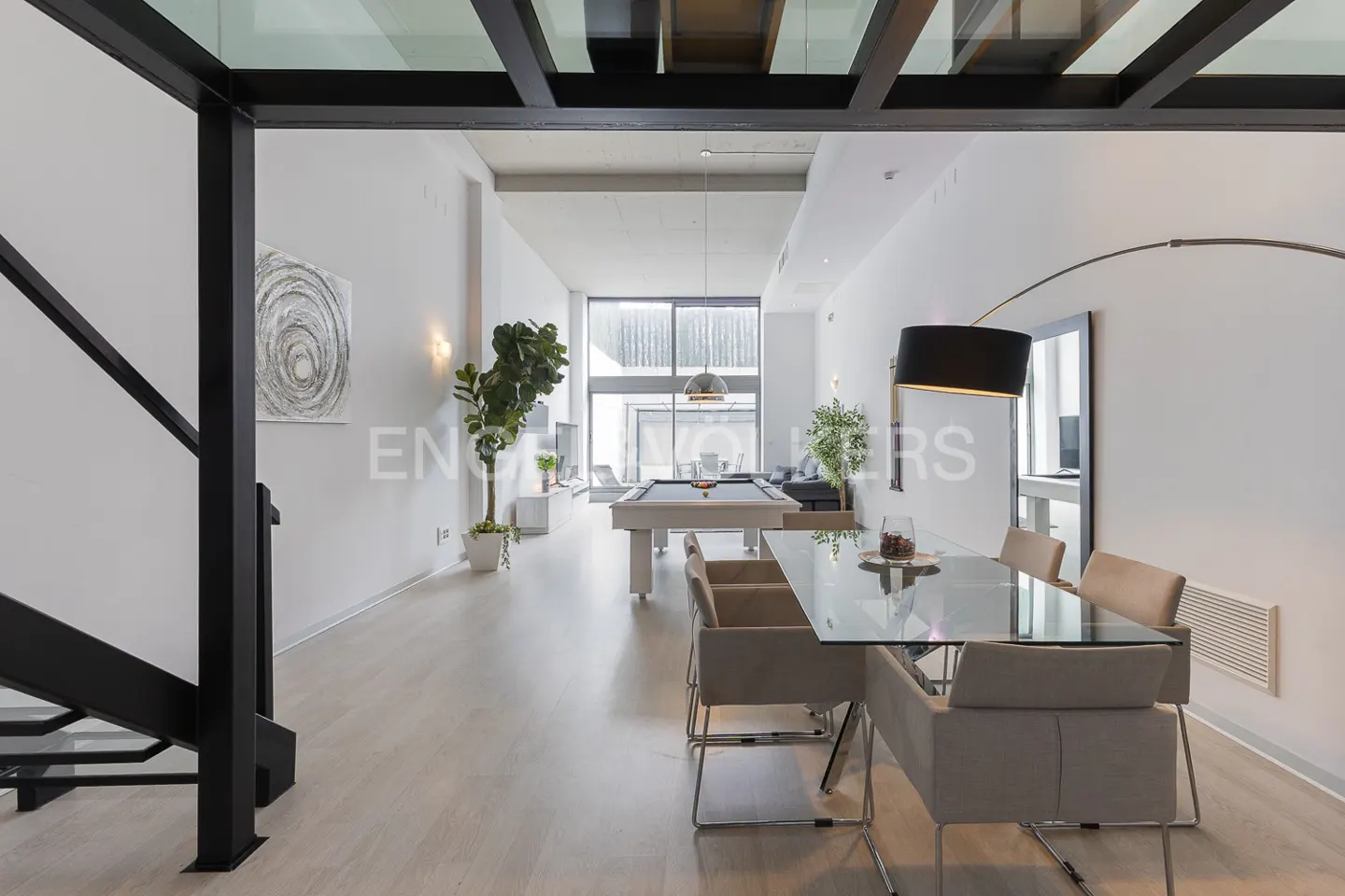Bright, modern interior with a glass dining table, pool table, and black staircase. White walls and light wood floors create a spacious feel.