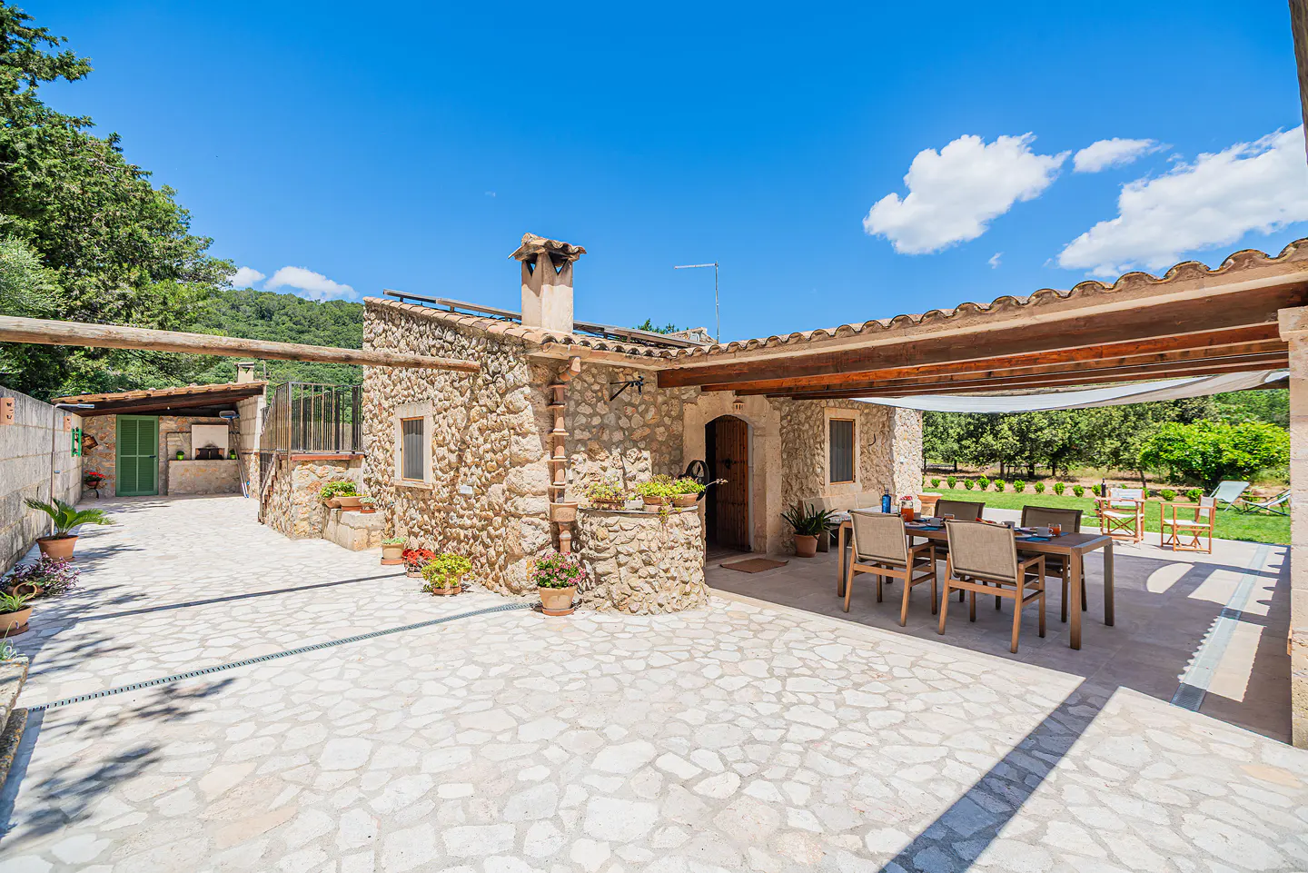 Stone house with patio, table, and chairs under a pergola. Blue sky and green trees in the background.