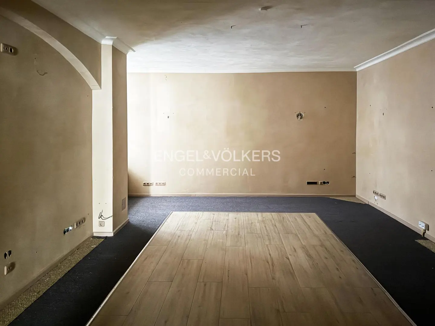 Unfurnished commercial space with beige walls, white trim, and mixed flooring of wood and dark carpet. Engel & Völkers logo on the wall.