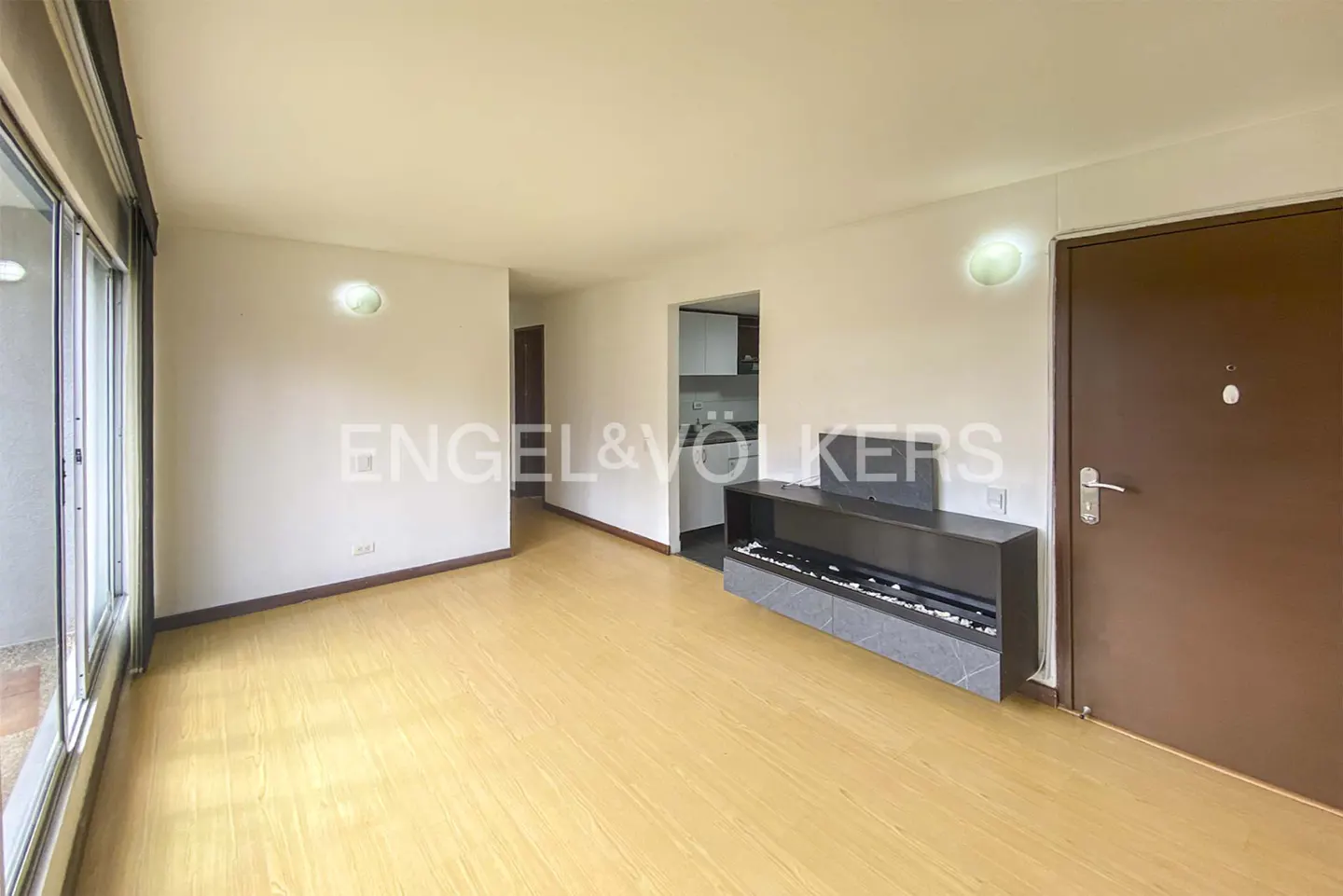 Bright living room with light wood floors, a modern wall-mounted fireplace, and a doorway leading to a kitchen.