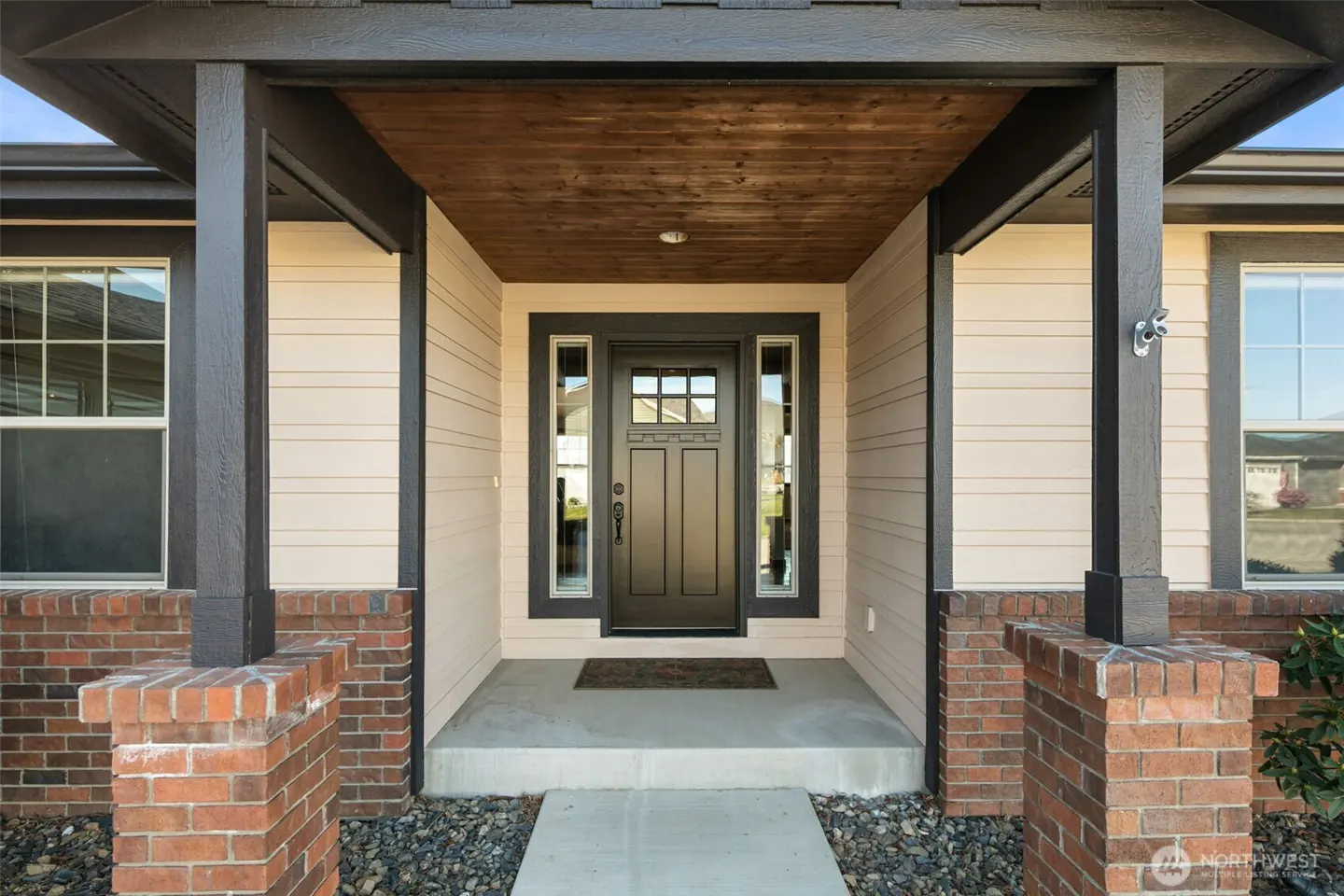Front entrance of a beige house with a dark brown door, brick pillars, and a wood-paneled porch ceiling.