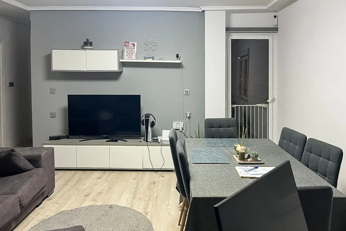 Living room with gray walls, light wood floors, and a gray dining table with chairs. A TV and console are against the wall.