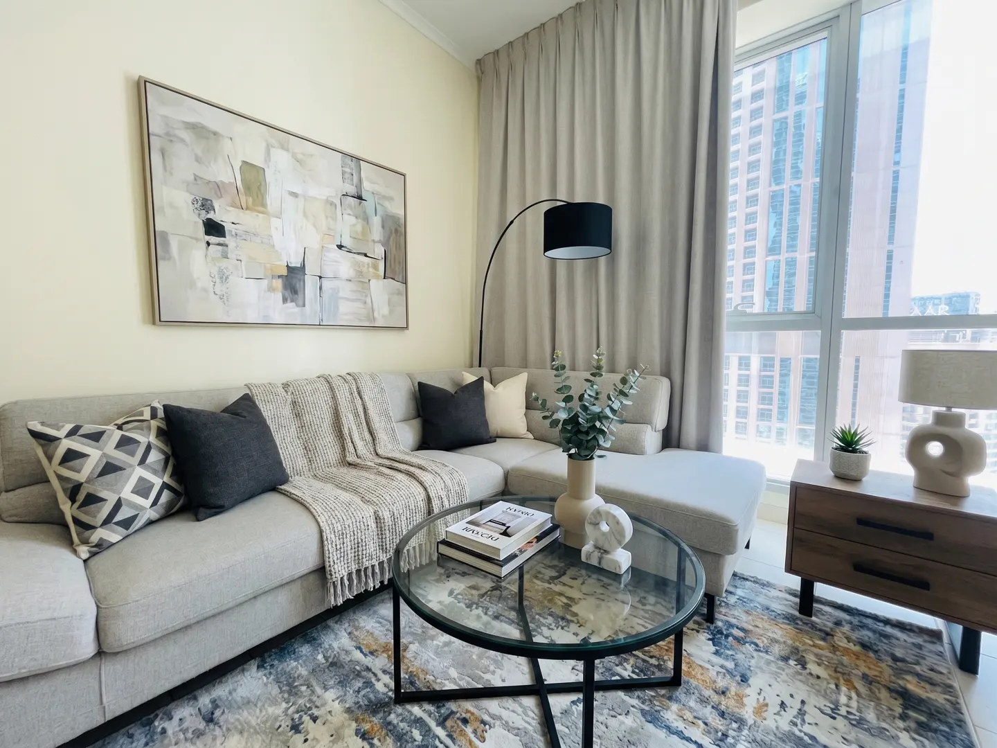 Living room with a gray sectional sofa, glass coffee table, and abstract art. Large window with city view. Neutral tones.