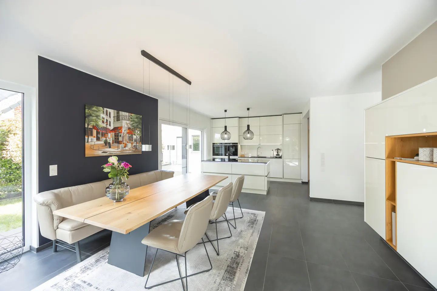 Open-concept kitchen and dining area with a long wooden table, bench seating, and modern white cabinets. A black accent wall features a colorful painting.