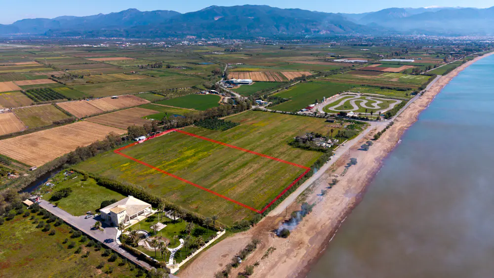 Aerial view of a green field outlined in red, next to a sandy beach and blue ocean, with mountains in the background.