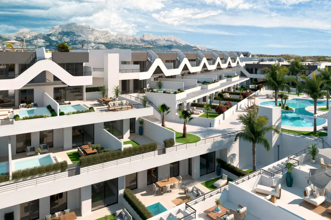 Aerial view of modern white condos with pools, patios, and palm trees under a blue sky with mountains in the background.