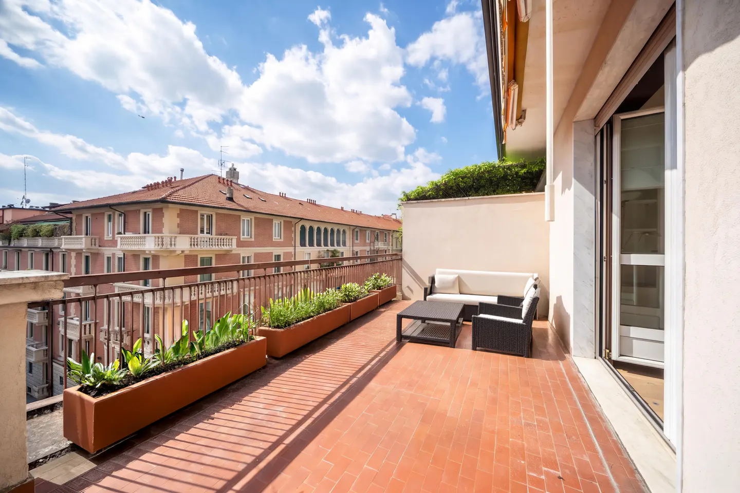 Balcony with terracotta tiles, planters, and outdoor furniture. A building is visible in the background under a blue sky.