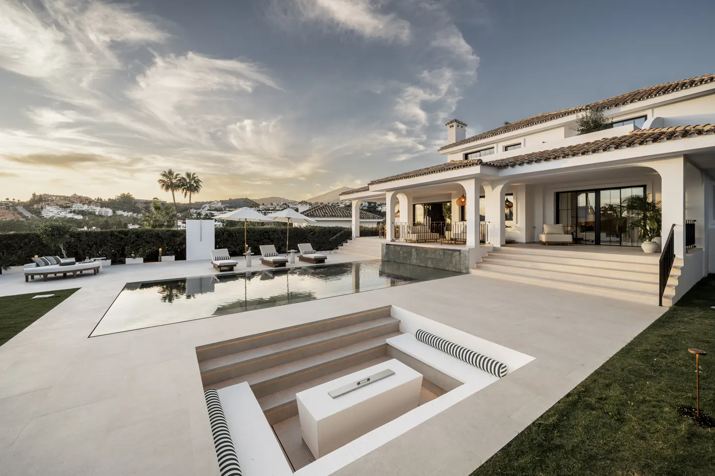 Luxury villa with a pool, lounge chairs, and a sunken seating area with a fire pit. The villa is white with a tile roof.