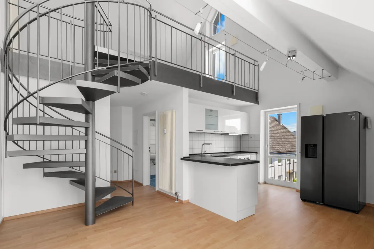 Modern apartment interior with a metal spiral staircase, white kitchen, black refrigerator, and a lofted mezzanine level.