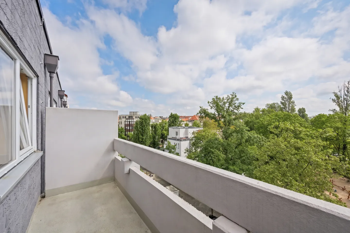 View from a gray balcony with a white railing, overlooking green trees and buildings under a cloudy blue sky.