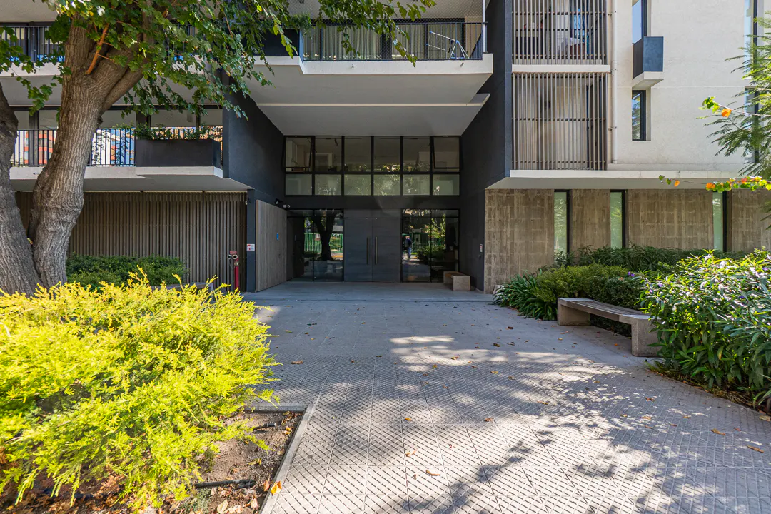 Apartment building entrance with glass doors, concrete walkway, and green bushes. Balconies are visible on the upper floors.