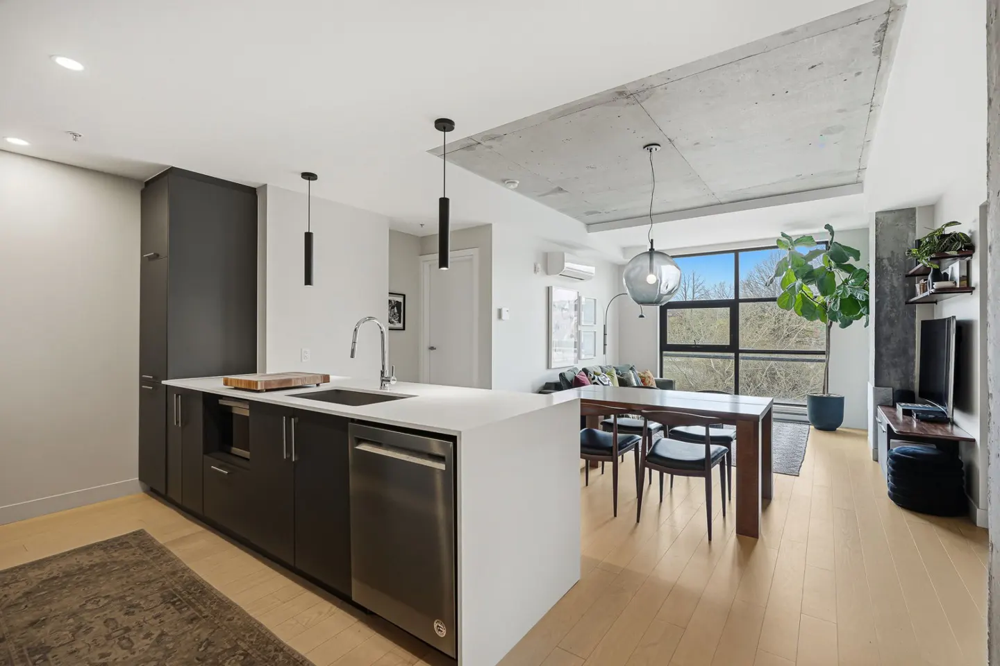 Modern apartment interior with an open floor plan. Kitchen island, dining table, and living area are visible. Large windows offer natural light.