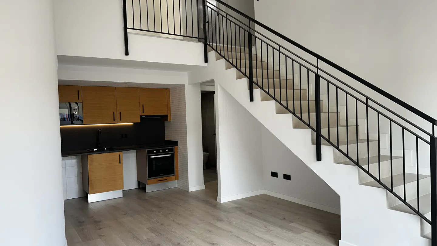Interior view of a modern apartment with a staircase, kitchen, and light wood floors. Black metal railings line the stairs and loft.