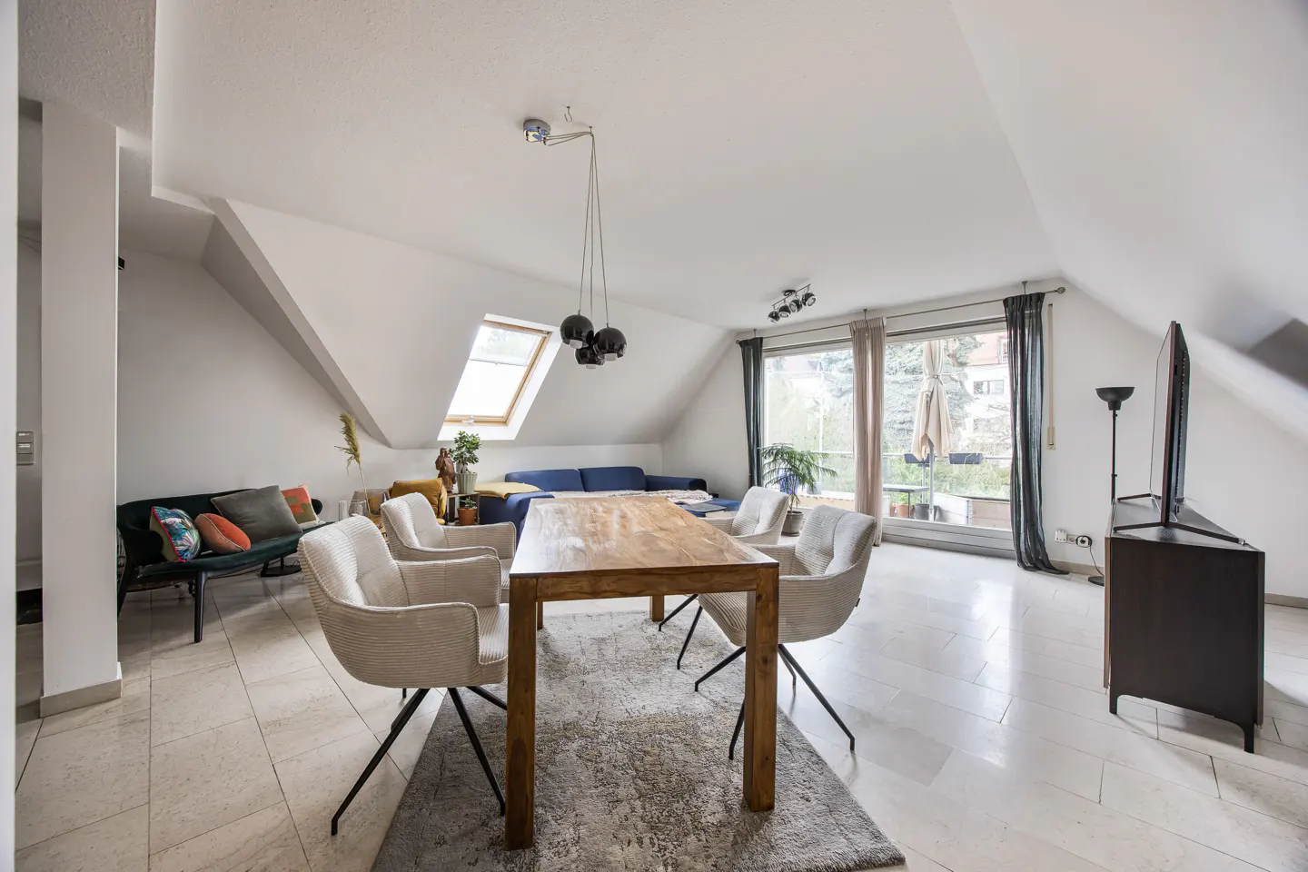 Bright, open attic apartment with a wooden dining table, white chairs, and a blue sofa. Large windows and a skylight provide natural light.