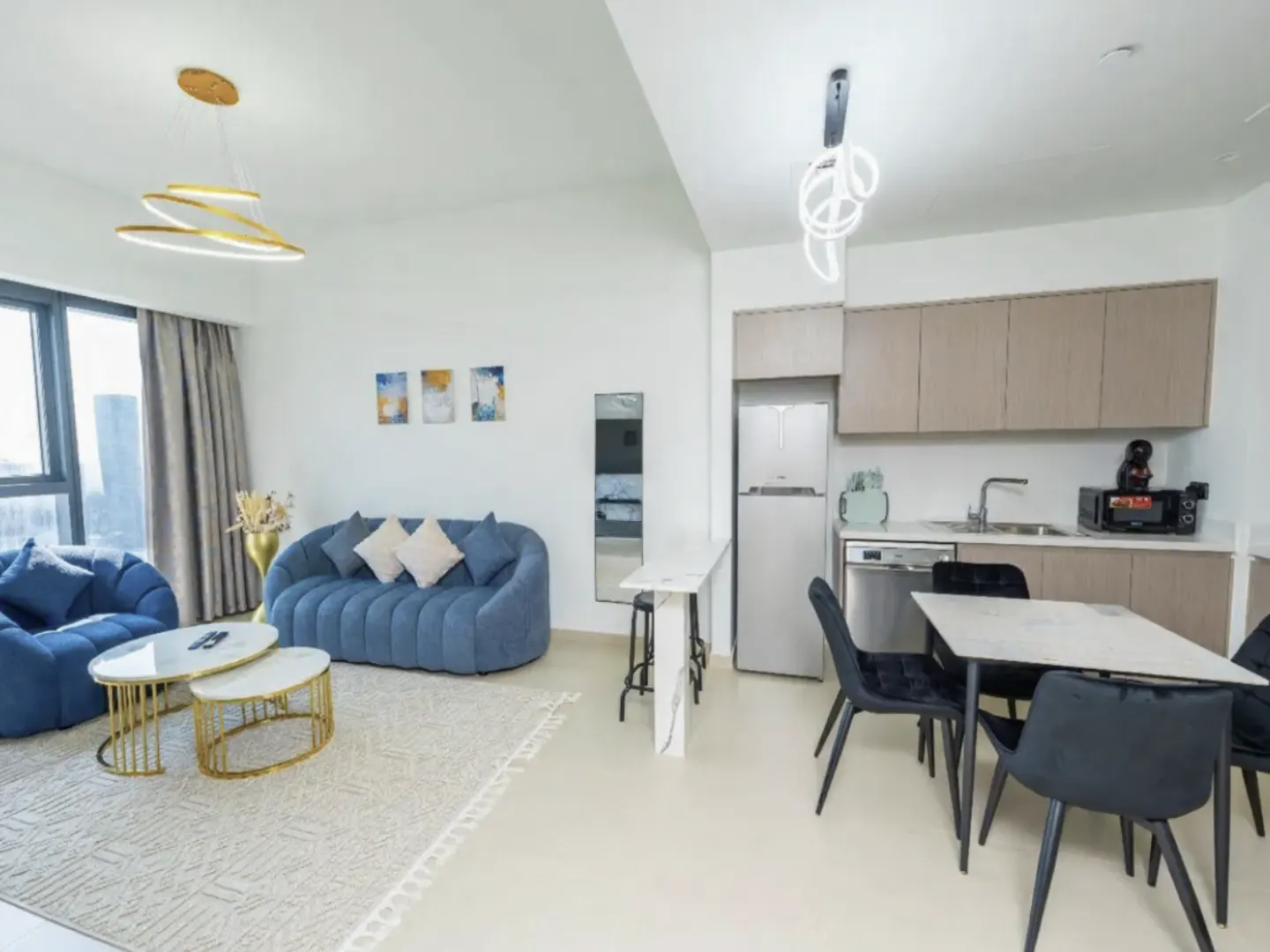 Modern open-plan apartment with a blue curved sofa, gold-accented coffee tables, dining area, and light wood cabinetry.
