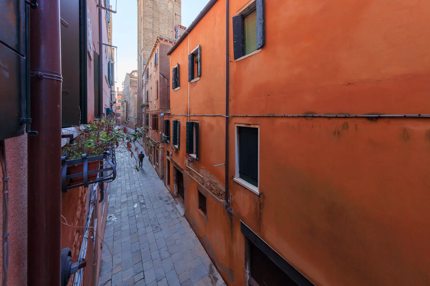A narrow, cobblestone street in Venice, Italy, lined with tall, orange buildings and dark green shutters. A person walks in the distance.